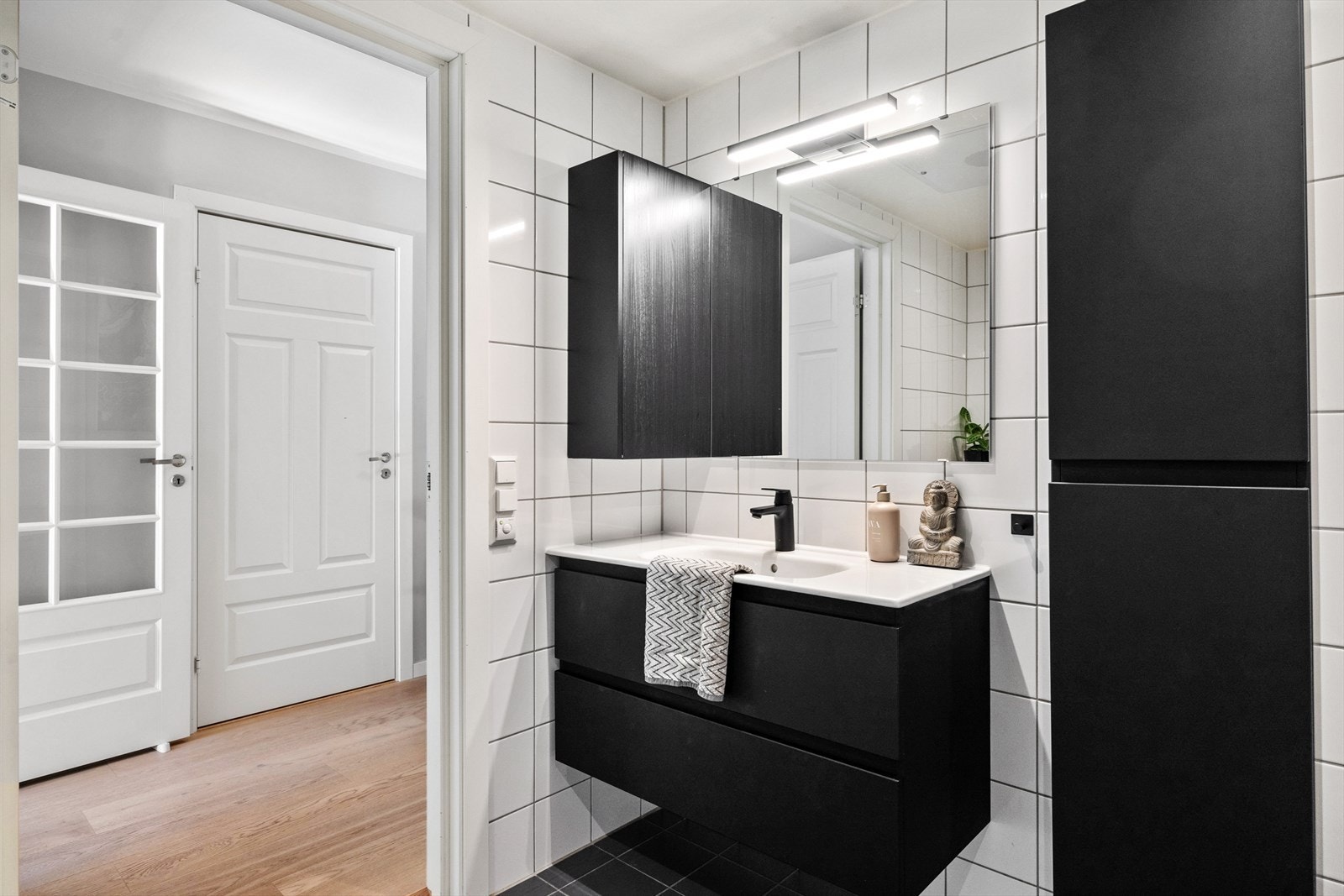 Badet består av servantskap med nedfelt servant, veggskap, speil med lys over, vegghengt toalett med innebygd sisterne, og dusjsone med innfelte dusjdører i herdet glass. Det er opplegg for vaskemaskin. Galleribilde