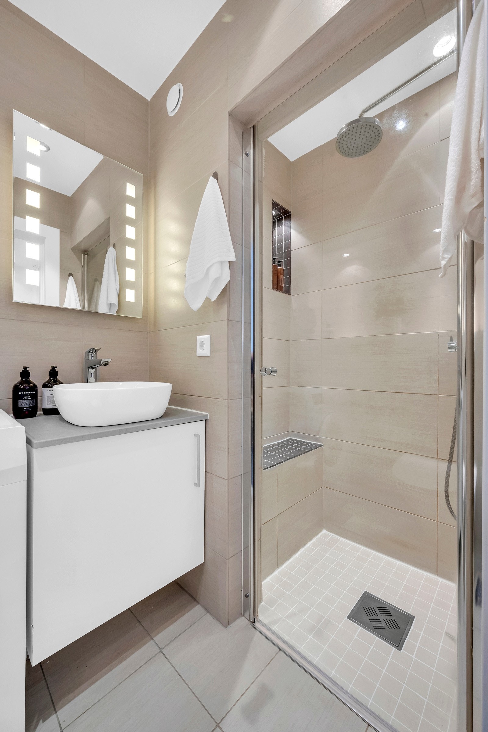 Badet er innredet med veggmontert klosett, toppmontert vask med innredning og speil og nedsenket dusjnisje med glassdører. Galleribilde