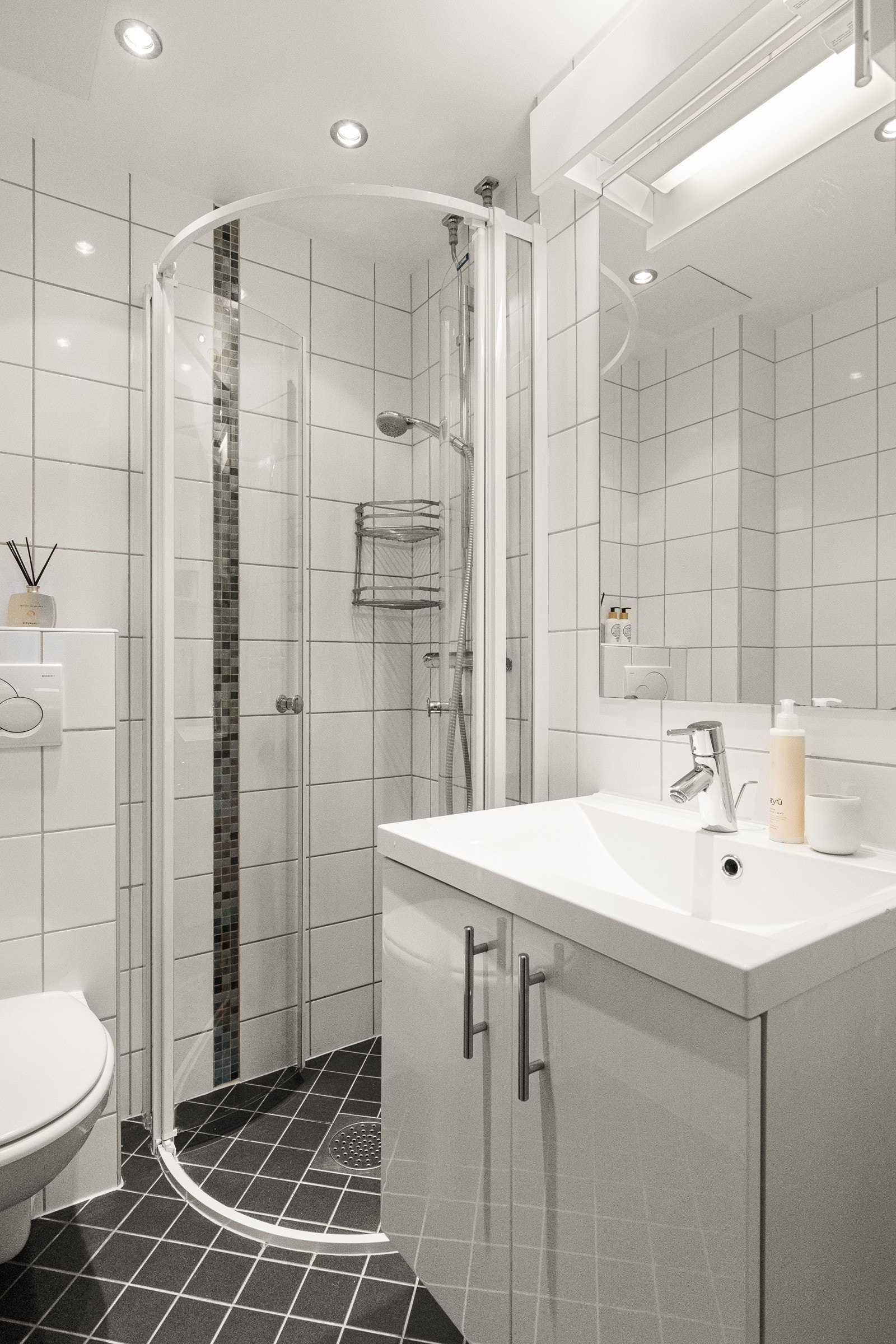 Baderommet inneholder servantskap og toalettskap med glatte høyglans fronter, speil og lysarmatur på vegg over servant, samt et veggmontert klosett. Galleribilde