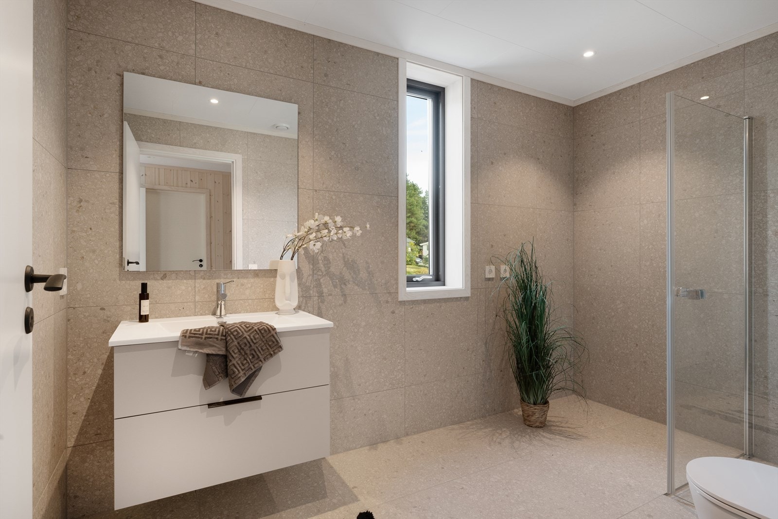 Badene består av 60x60 flislagte vegger og gulv med varmekabler. Flisene er av type Match Up Suger mix. Himling i tak-ess. Komplette baderomsmøbler fra Aubo som er levert i samme overflater som kjøkken. Galleribilde