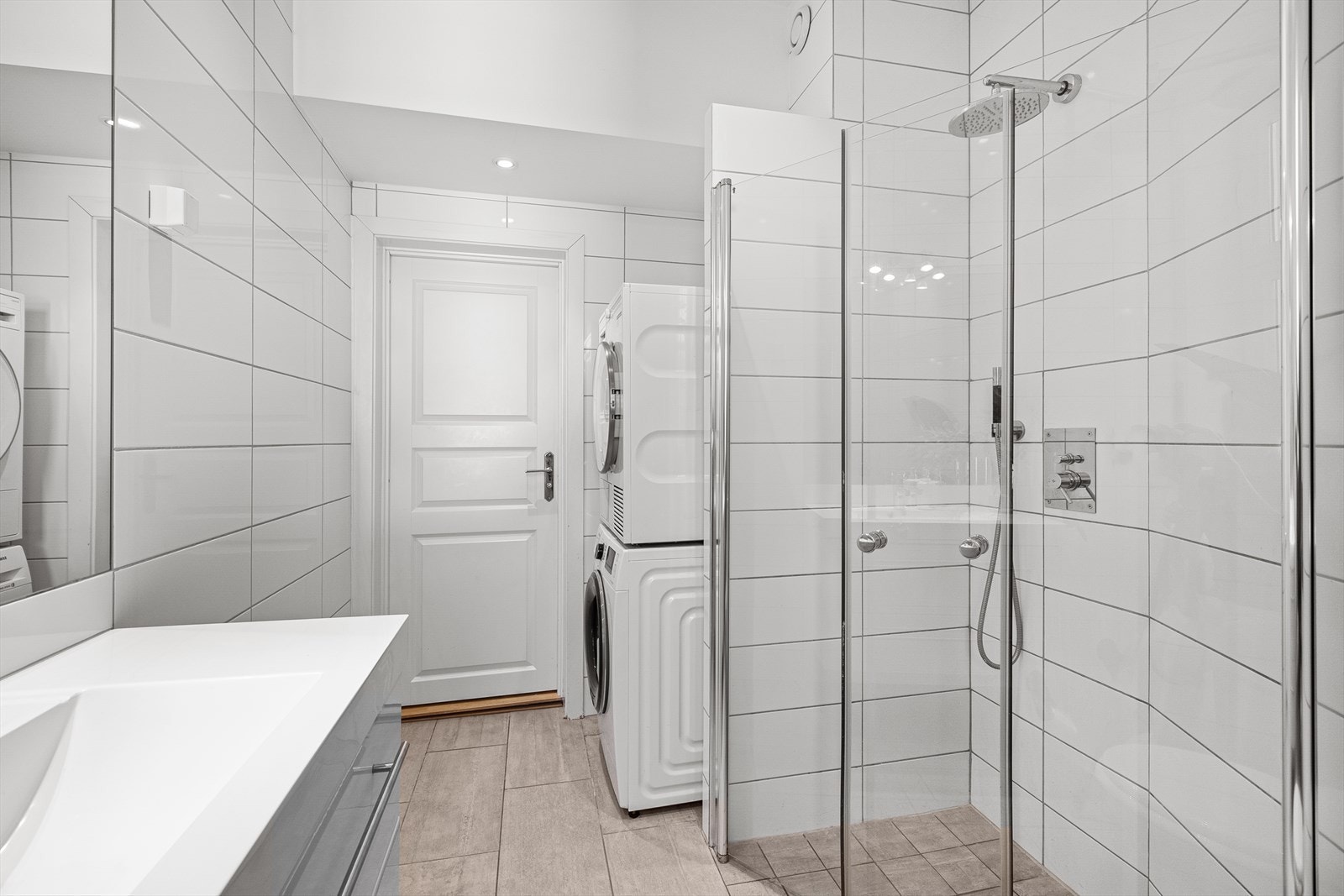 Baderommet inneholder veggmontert toalett, dusjhjørne med innfellbare glassdører og baderomsinnredning med skuffer servant og speil med belysning Galleribilde