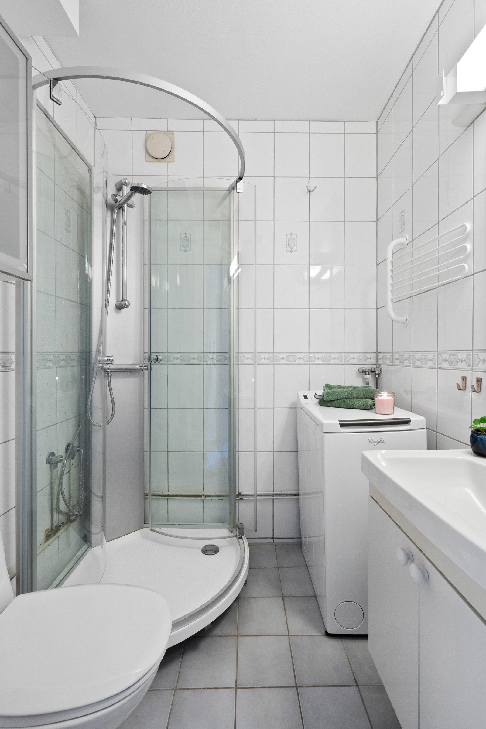 Badet i leiligheten har lysegrå gulvfliser og hvite veggfliser. Badet varmes opp med varmeelement med justerbar effekt over dør på badet. Galleribilde