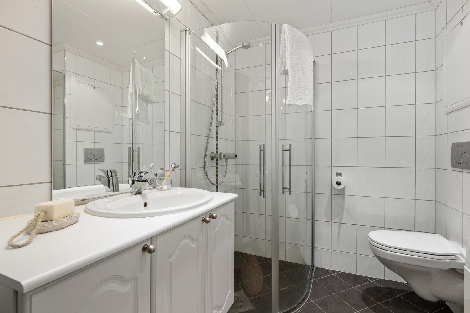 Badet er lyst i tidsriktige farger og kan skilte med vegghengt toalett, baderomsmøblement, dusj med glassdører samt opplegg til vaskemaskin. Galleribilde