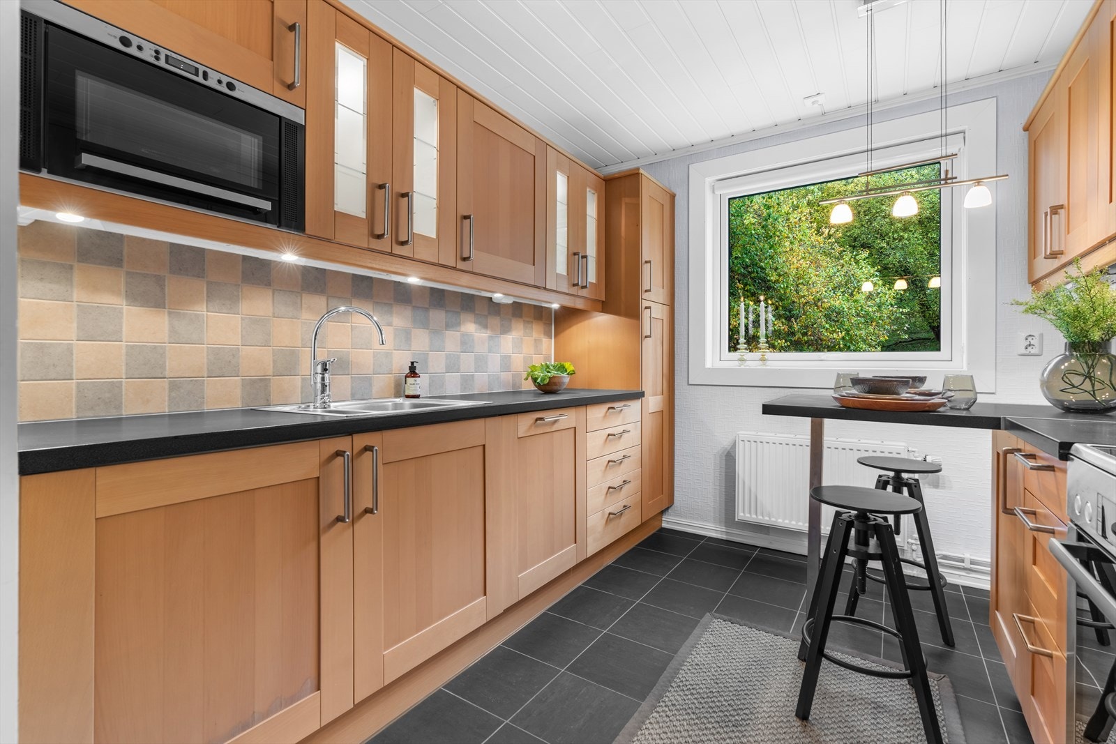 IKEA kjøkkeninnredning med profilerte fronter, enkelte glassfronter i overskap, belysning under overskap, fliser på vegg over benkeskap, laminat benkeplate med nedfelt kum i rustfritt stål og ventilator. Galleribilde