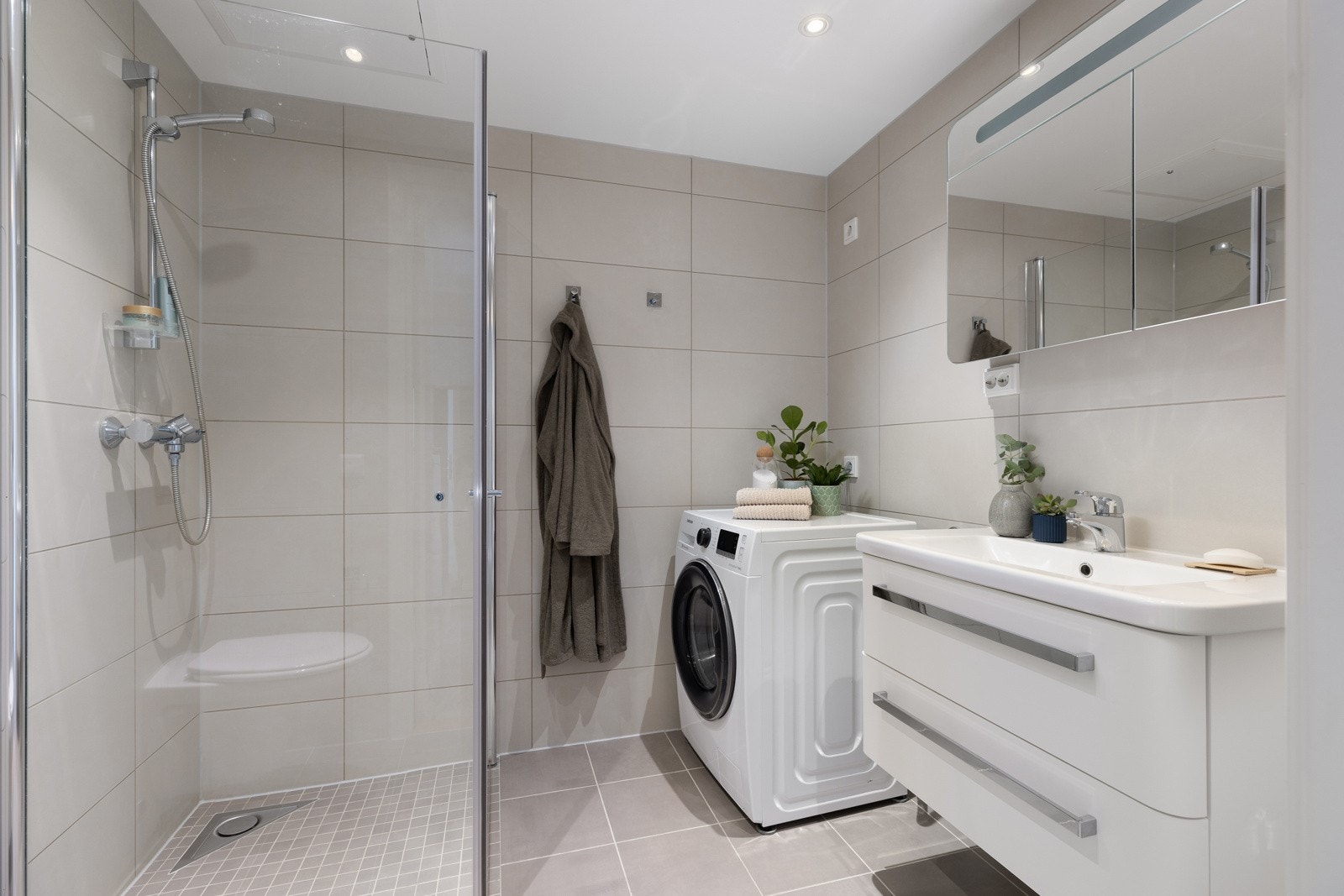 Moderne bad med flislagt gulv med varme, lyse veggfliser og downlights i himlingen. På badet er det opplegg for vaskemaskin. Galleribilde