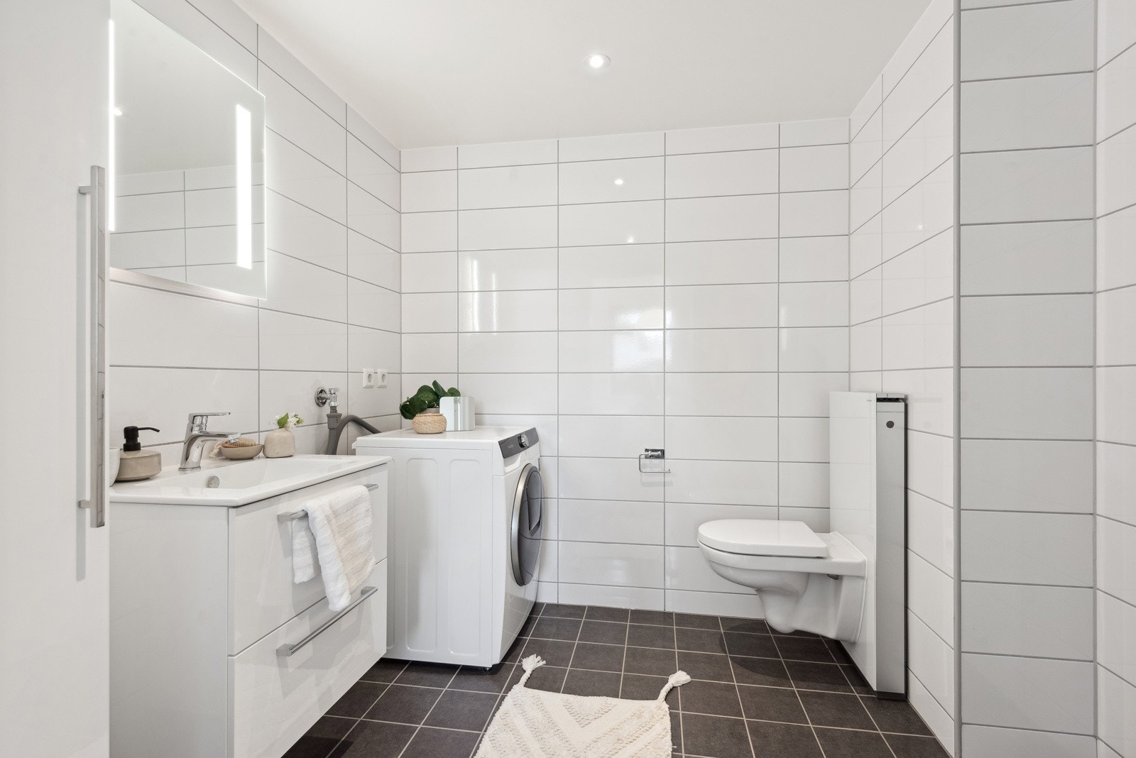 Badet har lyse og moderne overflater. Det er vegghengt toalett, dusjnisje og servant med skuffer under. Det er også opplegg for vaskemaskin og tørketrommel. Galleribilde