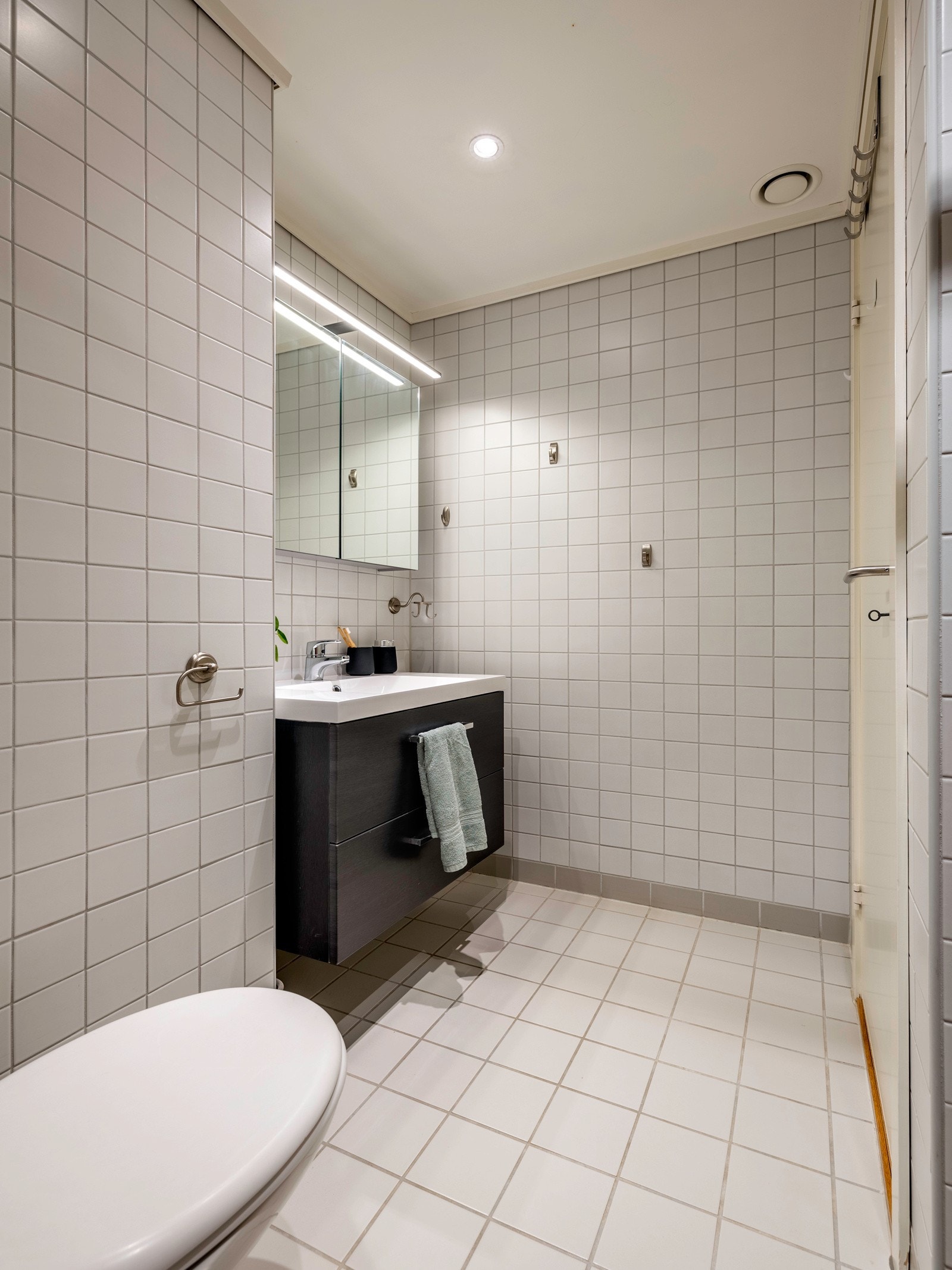 Badet er utstyrt med et gulvstående toalett, dusjhjørne med forheng, og en servant med ettgreps blandebatteri Galleribilde