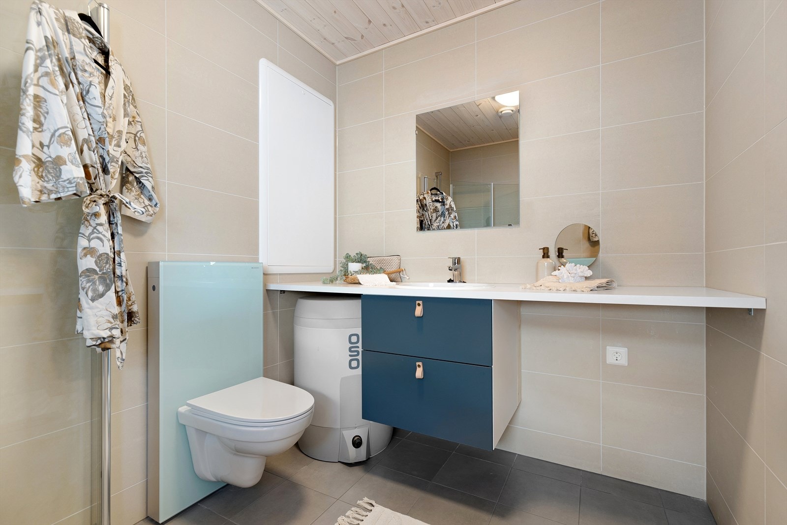 Badet er enkelt og minimalistisk innredet, med vegghengt toalett, antrasittgrå fliser på gulvet og beige fliser på vegg. Galleribilde