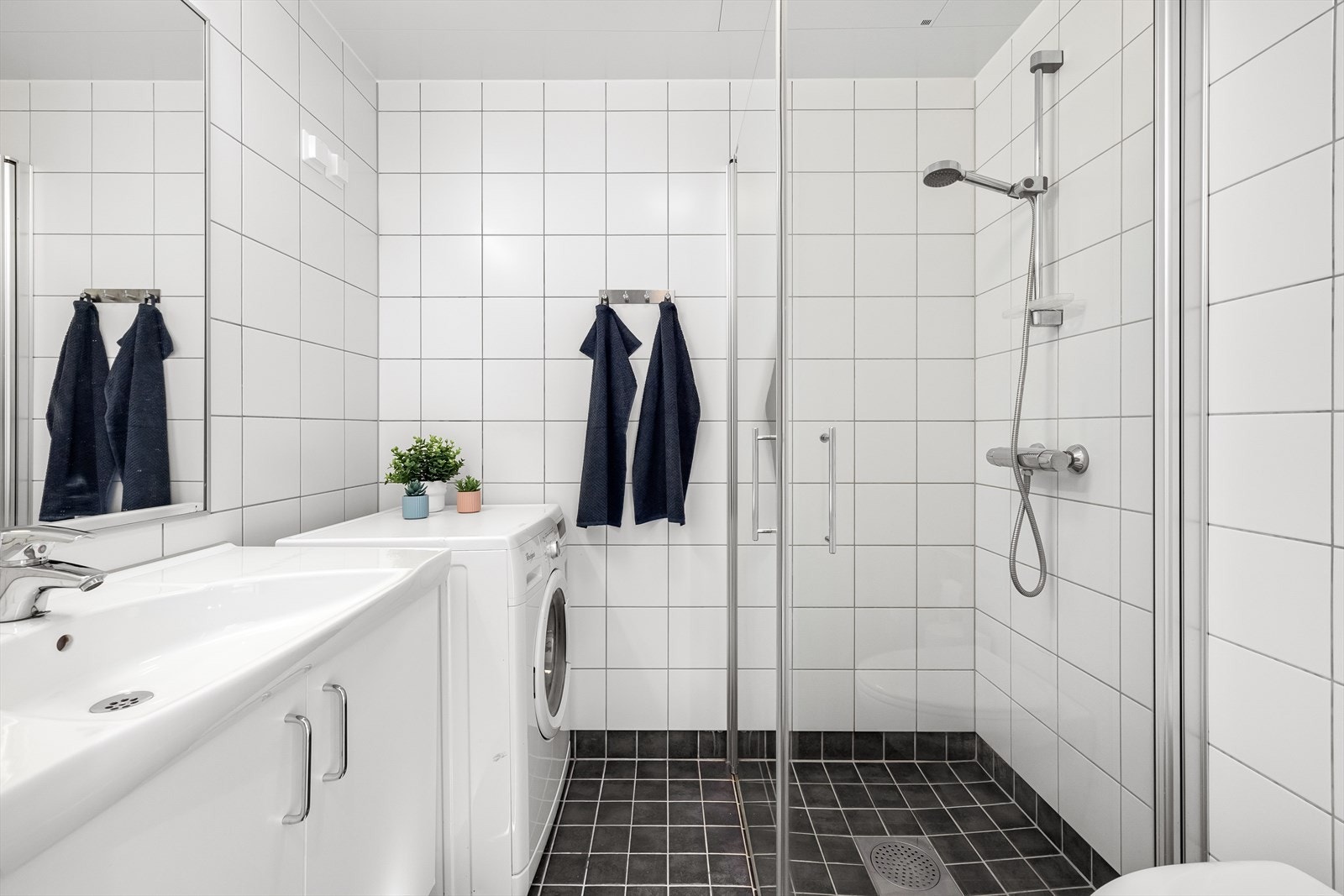 Badet er utstyrt med dusjhjørne med innfellbare dusjdører med blandebatteri fra Oras, veggmontert toalett og baderomsinnredning med servantskap, hvit servant og bladebatteri fra Oras. Galleribilde