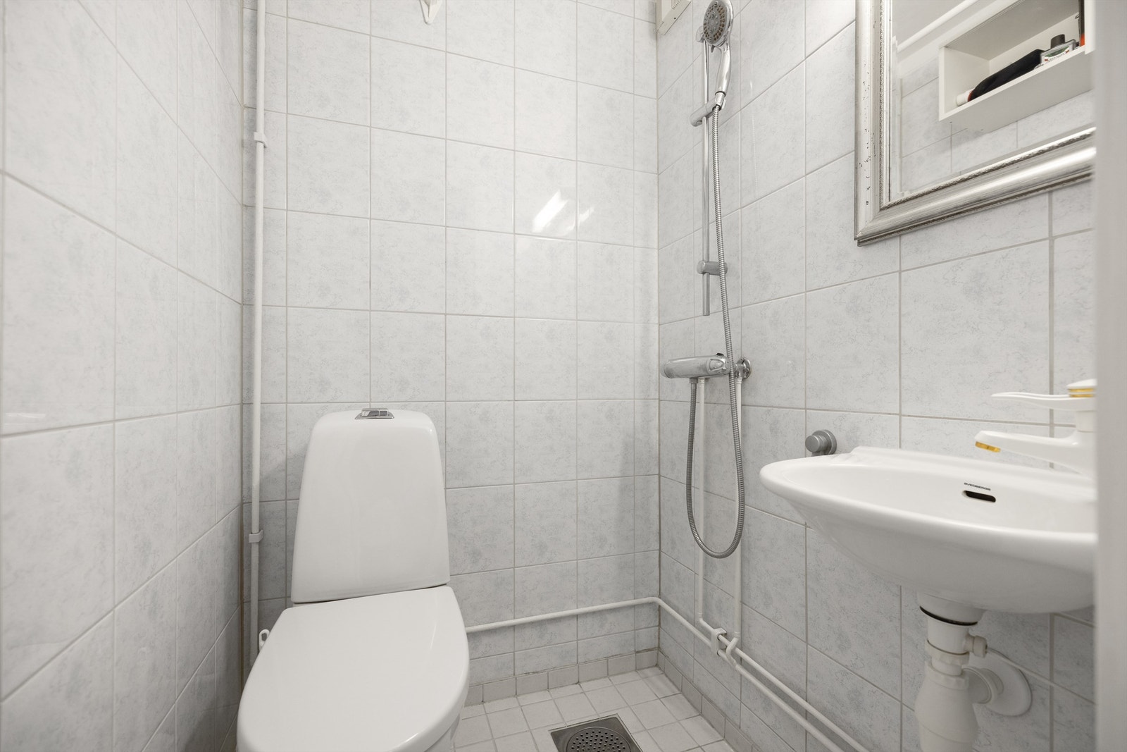 Eldre og flislagt bad med gulvvarme. Badet består av vegghengt servant med armatur, speil, overlys over servant, dusjhjørne med forheng, dusjarmatur tilkoblet hånddusj og gulvstående toalett Galleribilde