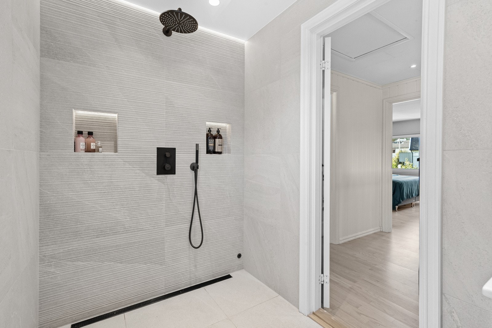 Badet er luksuriøst med skjulte rør i veggen for et stilrent uttrykk. Det har en bred dusjnisje med veggmontert regnfallsdusj, hånddusj, sort kran og slukrenne, samt to innfelte veggnisjer med belysning. Galleribilde