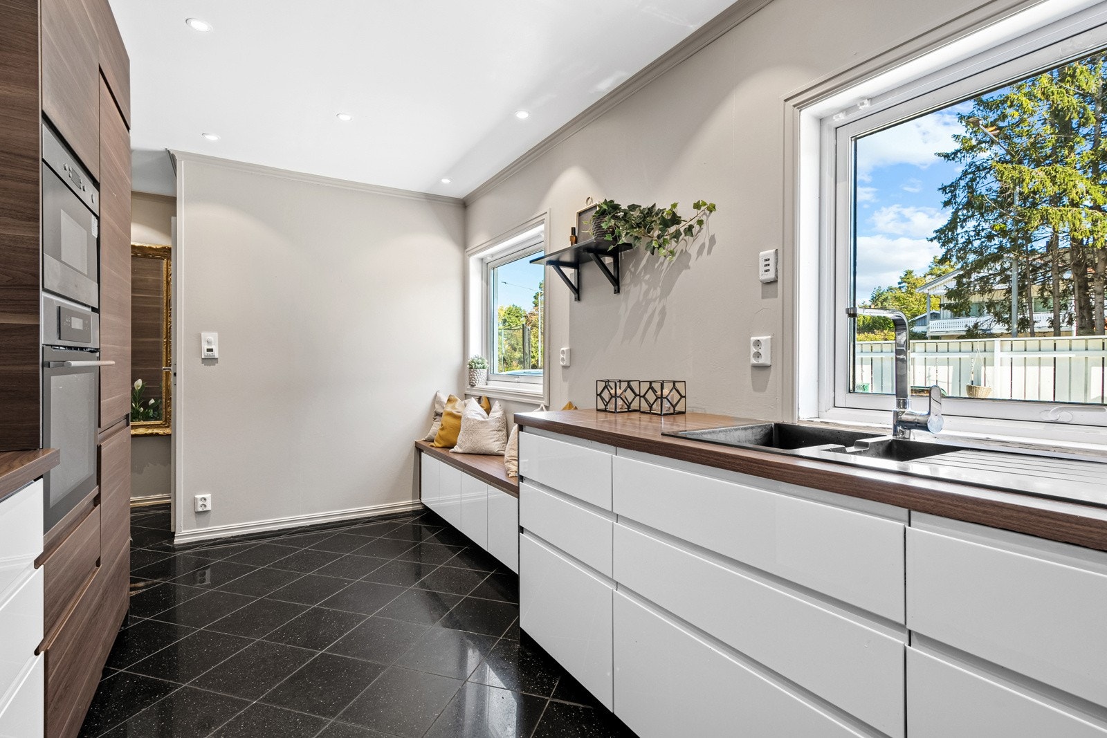 Rommet har sorte, polerte gulvfliser som flyter sømløst inn i trapperom og entré. Benkeskapene har stilrene hvite fronter, og benken er utstyrt med en nedfelt vaskekum i mørk kompositt. Galleribilde