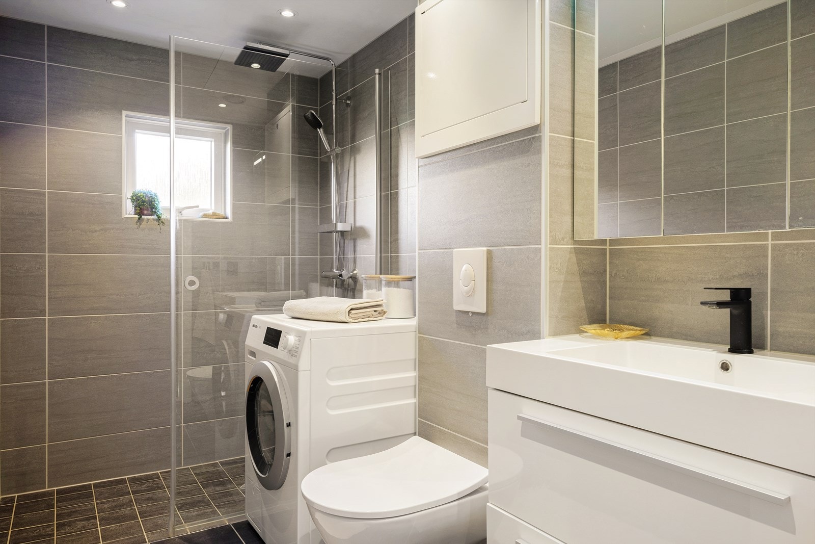 Lyst og helfliset bad med varmekabler i gulvet. Badet er utstyrt med vegghengt toalett, helstøpt servant, skuffer under servant, dusjdører i klart glass, opplegg for vaskemaskin og fordelerskap til rørsystemet Galleribilde