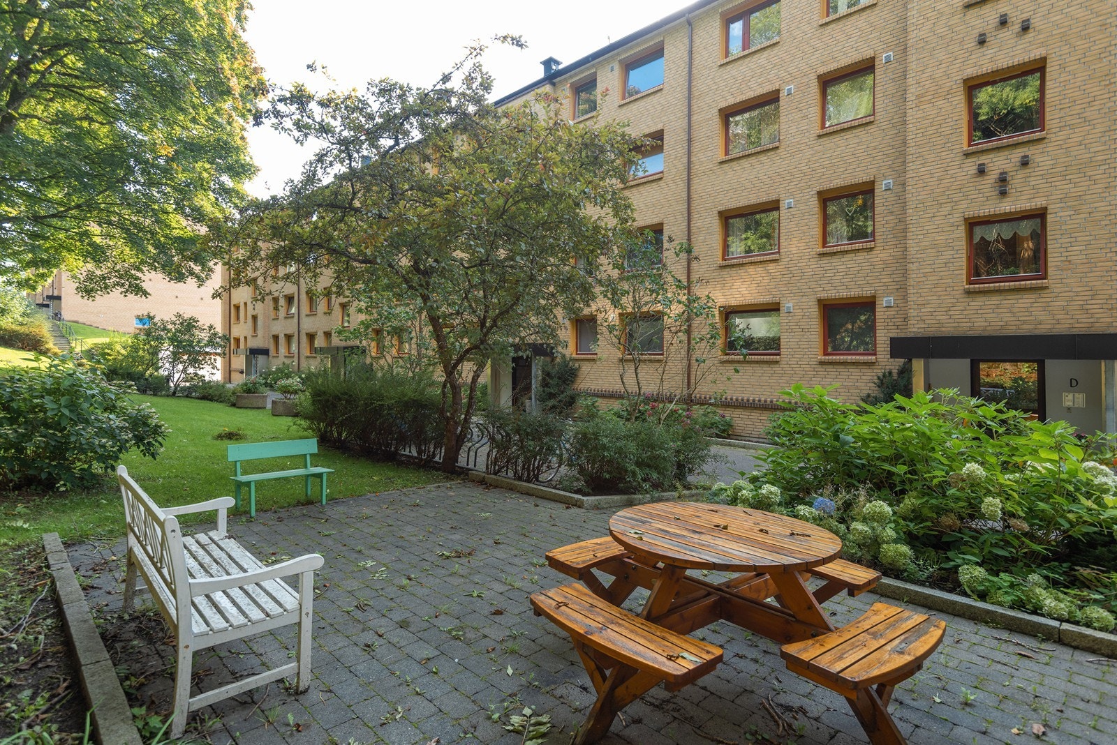 Ved inngangspartiene er det en felles uteplass med sittebenker blant bed, trær og div. grøntareal. Galleribilde