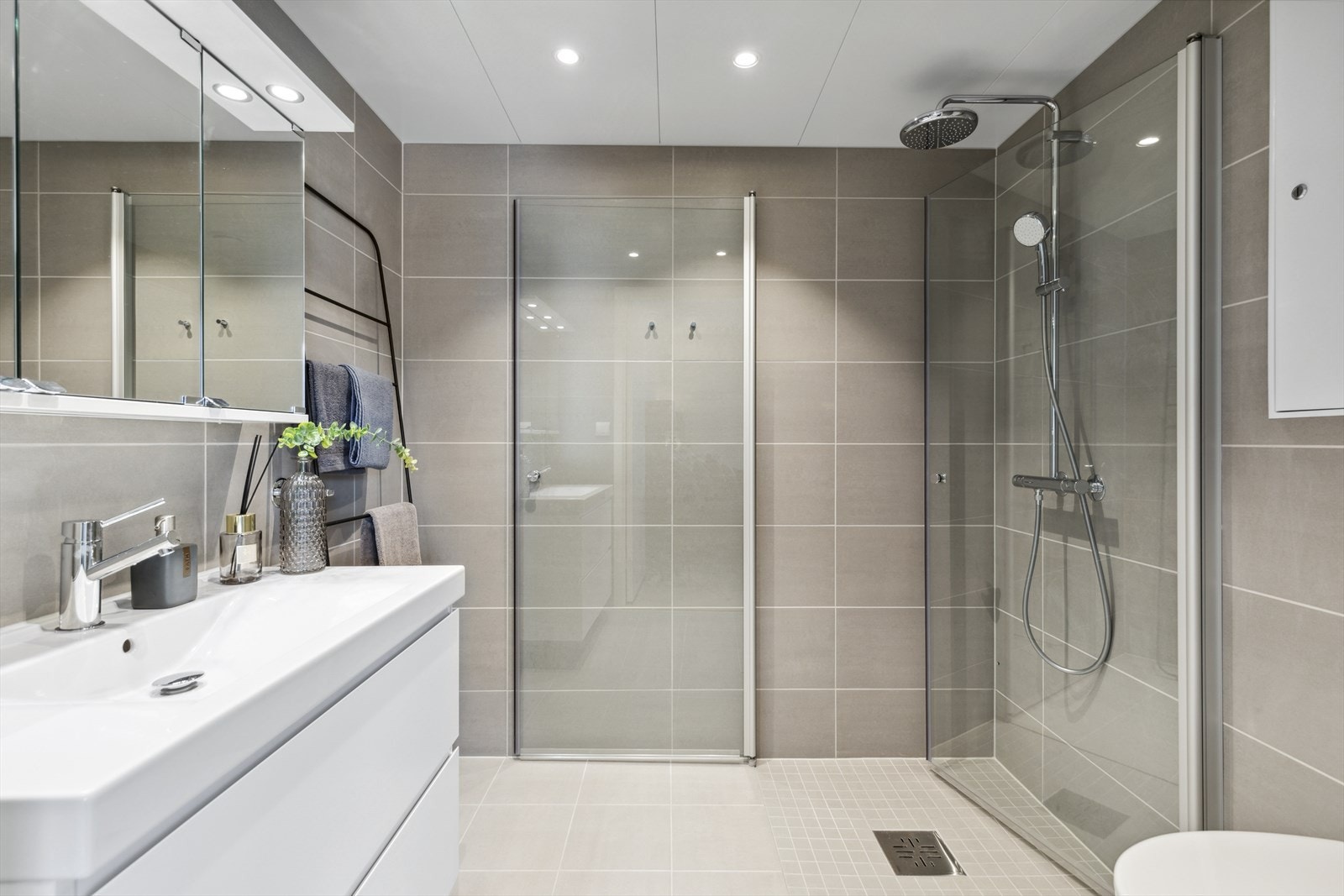Badet her har servant, veggmontert toalett, dusjvegger/hjørne og har delikate lysegrå fliser på vegger. Galleribilde