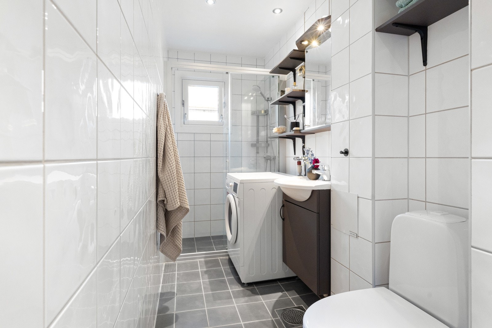 Fint, flislagt baderom som består av dusjhjørne, servant over innredning, speilskap med lys og toalett. Galleribilde