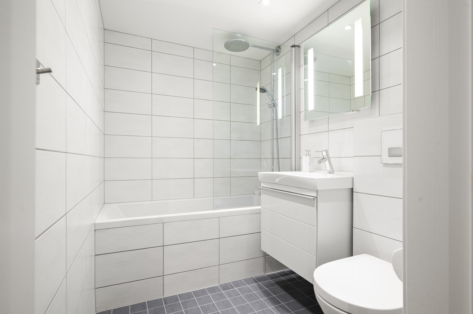 Badet er pent flislagt på gulv og vegger med varme i gulvet. Baderomsinnredningen består av vegghengt servantinnredning med servant. Badet har badekar med dusj. Galleribilde