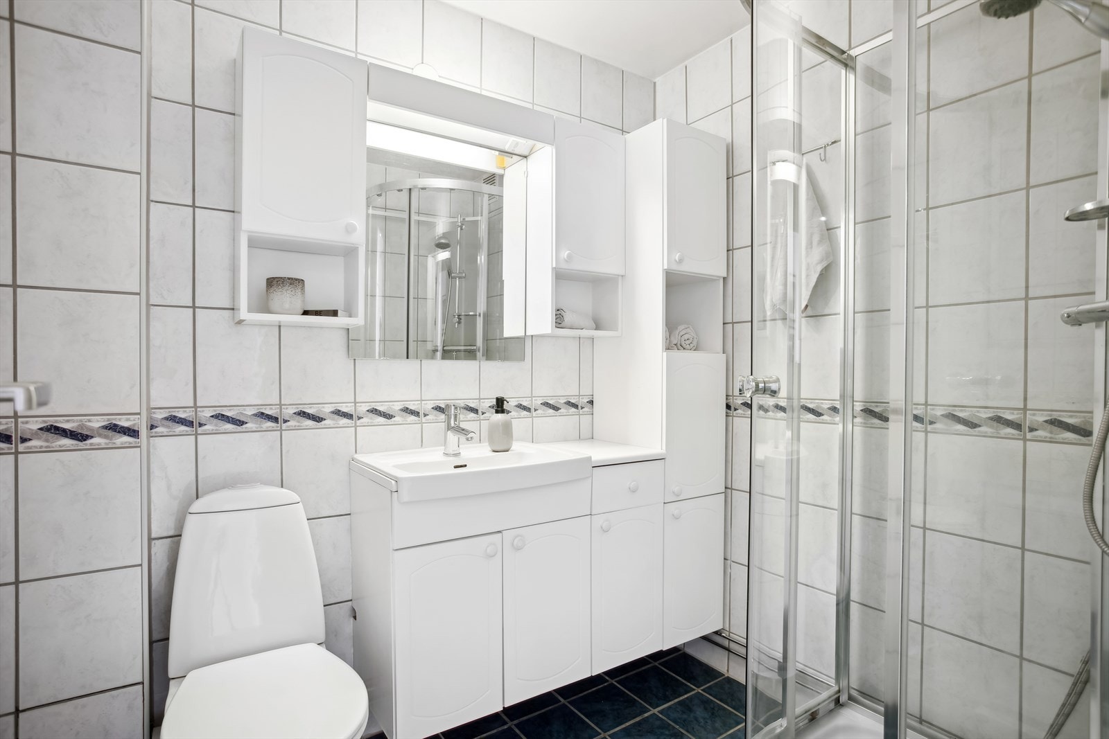 Badet er utstyrt med servantskap og høyskap, servant, speil over servant, dusjkabinett med 2 svingbare dusjvegger og gulvstående toalett. Galleribilde