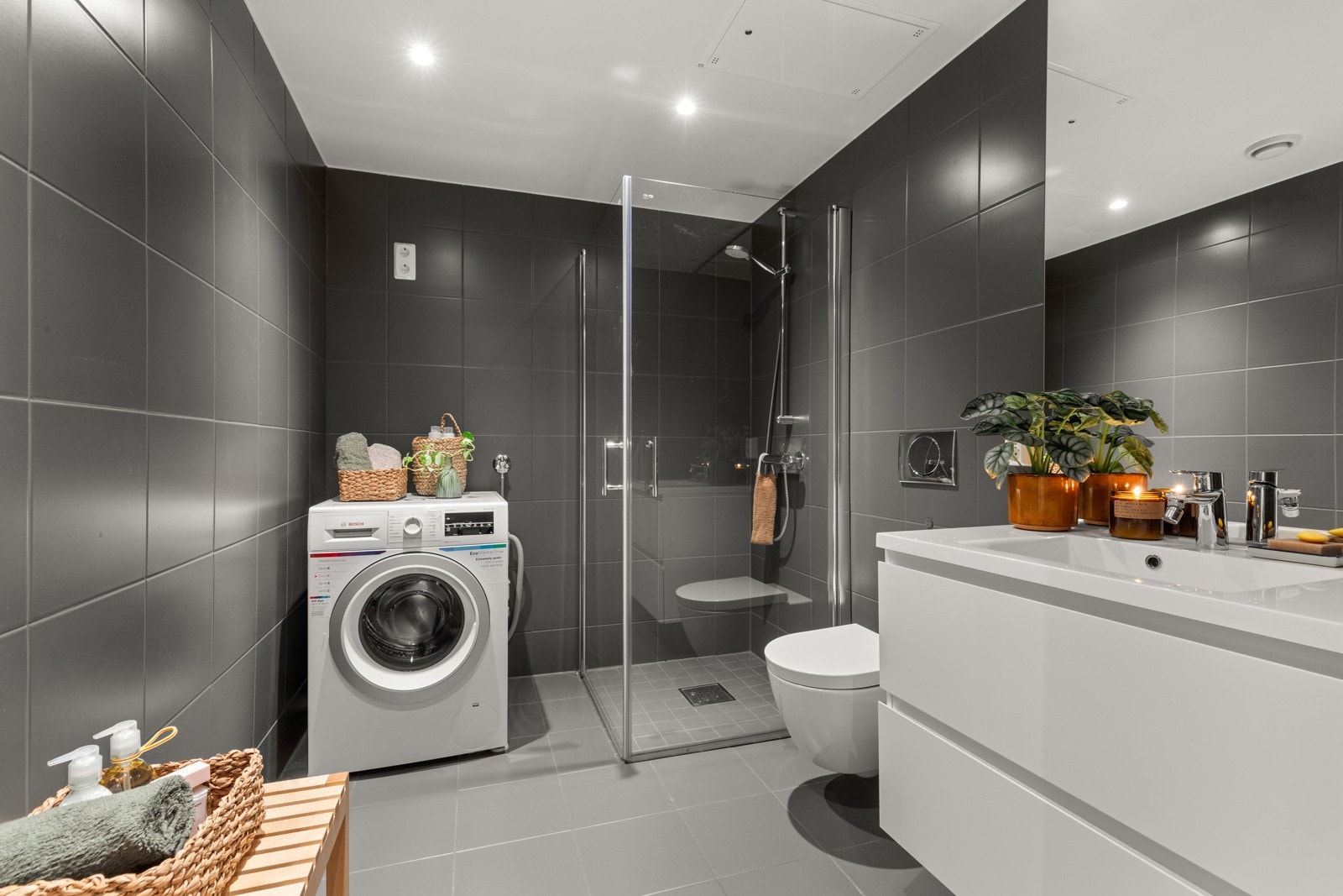 Badet er utstyrt med dusj med innfellbare glassvegger, servantseksjon med skuffer og heldekkende komposittvask, speil med lys, vegghengt wc og opplegg for vaskemaskin/tørketrommel. Galleribilde