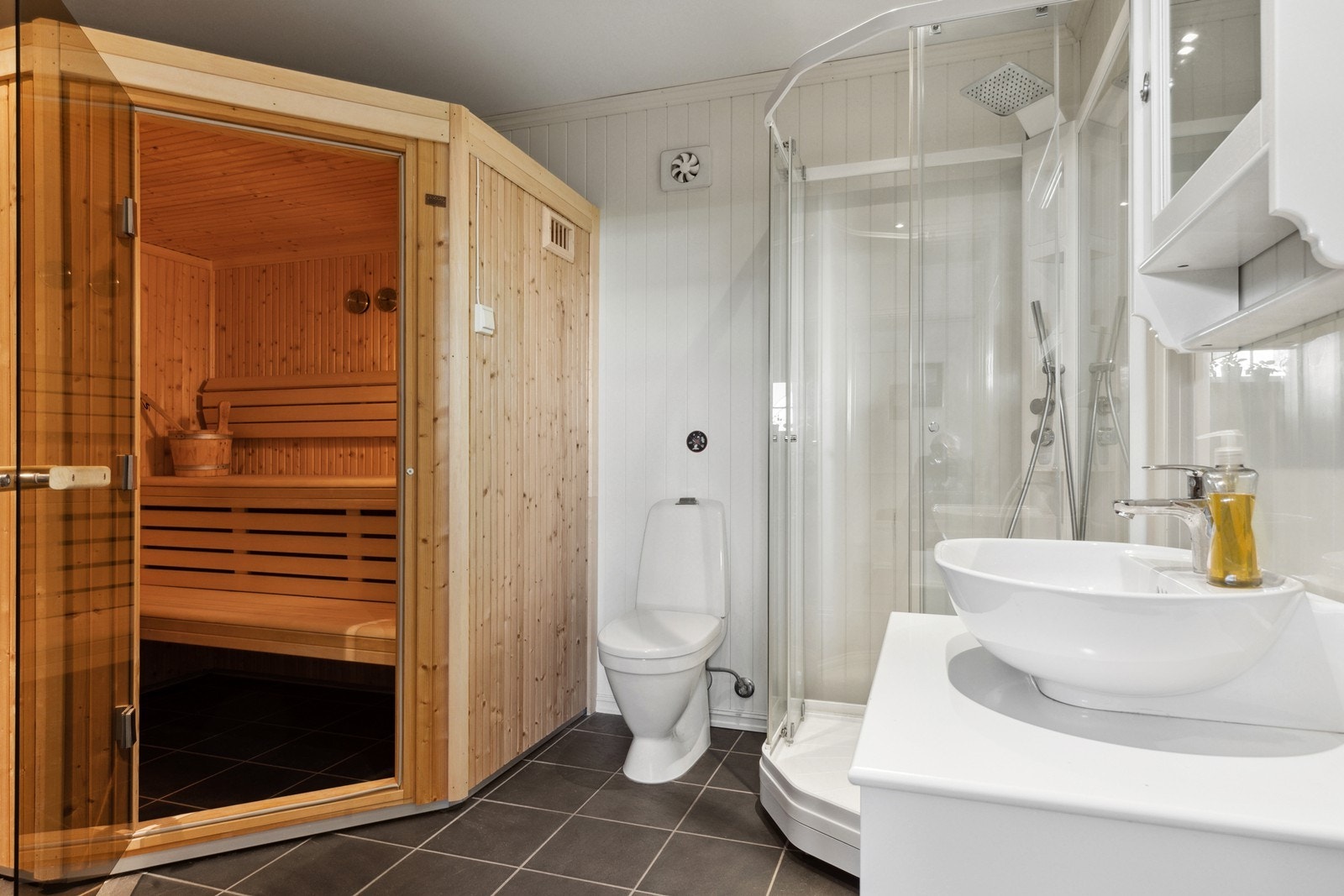 Bad med fliser på gulv med elektriske varmekabler og malte mdf-plater på veggene. Rommet er innredet med servantinnredning, toalett og dusjkabinett. Galleribilde