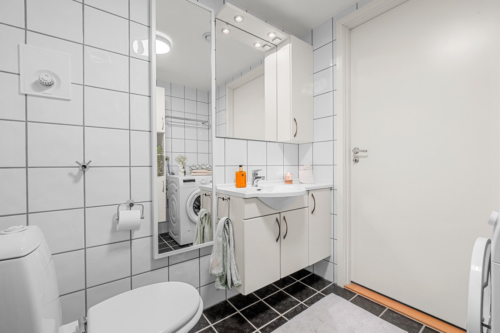 Baderommet innehar dusjdører av glass med dusjgarnityr på vegg, innredning med servant, gulvmontert toalett og opplegg for vaskemaskin. Galleribilde