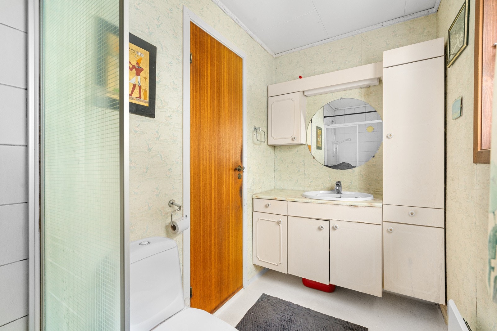 Eldre baderom med belegg på gulv og tapet/fliser på veggene. Dusjnisje med forheng, gulvstående toalett og naturlig ventilasjon. Galleribilde