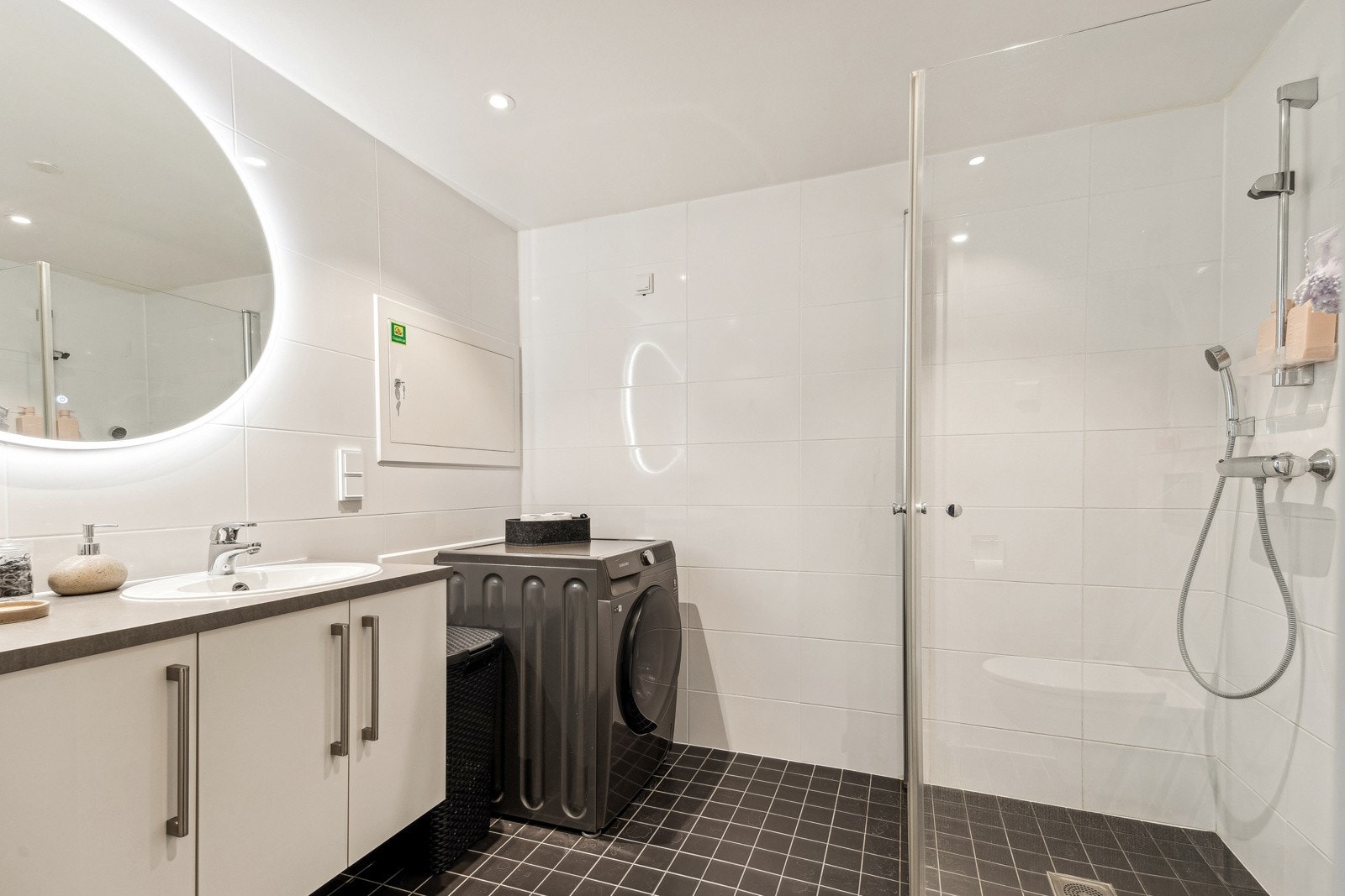 Lyst baderom utstyrt med vegghengt servantinnredning, speil med lys, dusjhjørne, vegghengt toalett og opplegg for vaskemaskin. Galleribilde