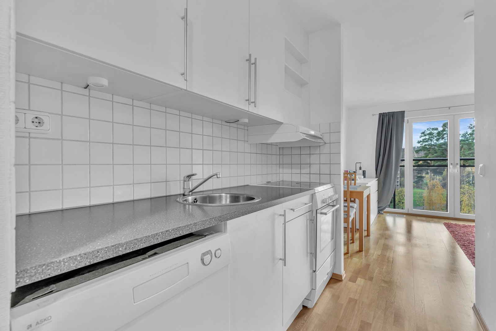 Videre på kjøkkenet er det fliser lagt mellom benkeplate og overskap, stikkontakter på vegg og belysning under overskap, frittstående komfyr med keramisk platetopp plassert i nisje. Det er ventilator med belysning og avtrekk ut av bygget, innebygget oppva Galleribilde