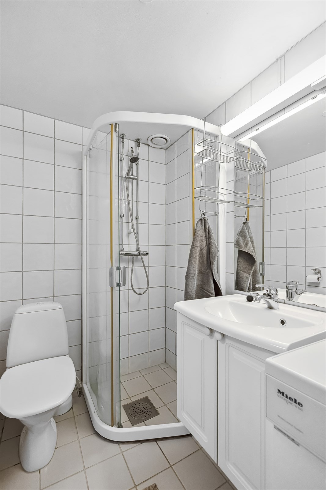 Badet er helfliset og har varmekabler i gulvet. Det er innredet med dusjhjørne, servant, speil med overlys, toalett og opplegg for vaskemaskin. Galleribilde