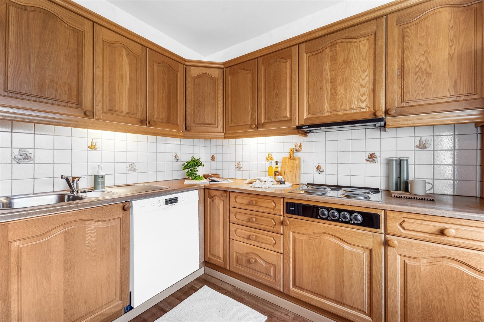 Kjøkkeninnredning med eikefronter, laminat benkeplate med fliser over, ventilator, opplegg for oppvaskmaskin og integrerte hvitevarer som stekeovn og koketopp Galleribilde