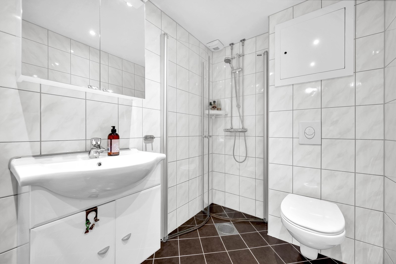 Badet har downlights i himling, dusjhjørne, opplegg for vaskemaskin, servant med oppbevaring, vegghengt toalett og varmekabler i gulv. Galleribilde