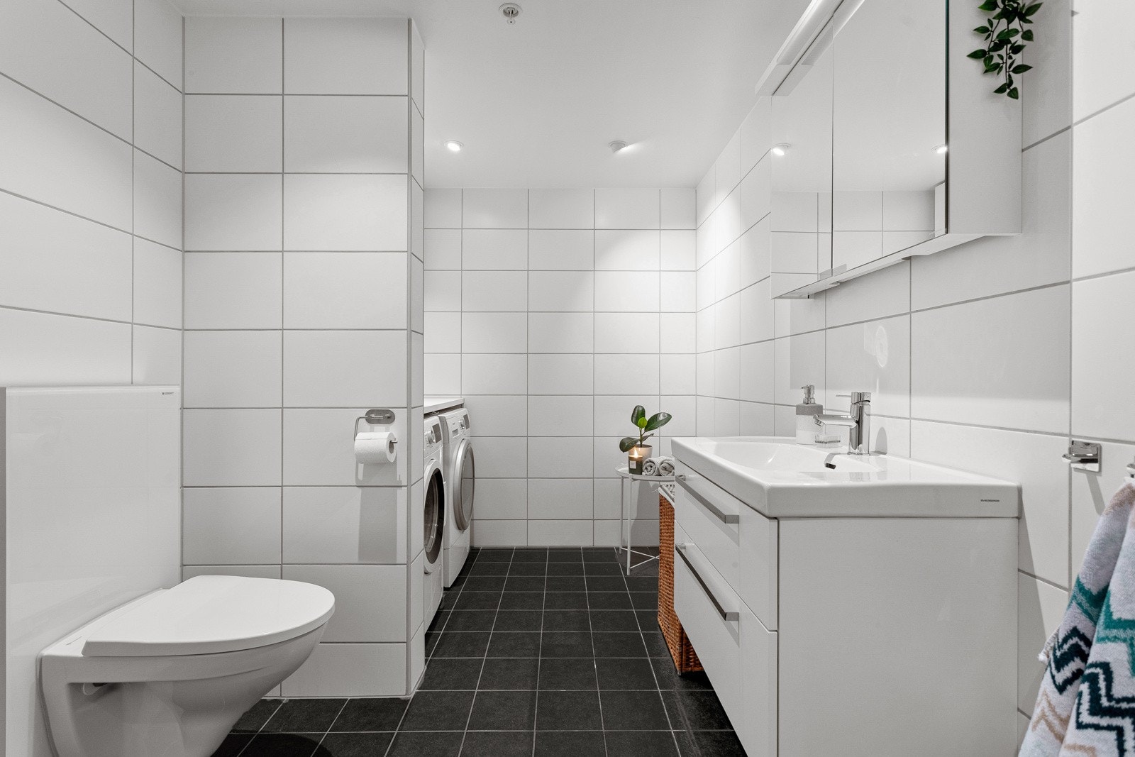 Baderommet har mørke fliser på gulv med gulvvarme, hvite fliser på veggene og downlights i himlingen. Galleribilde