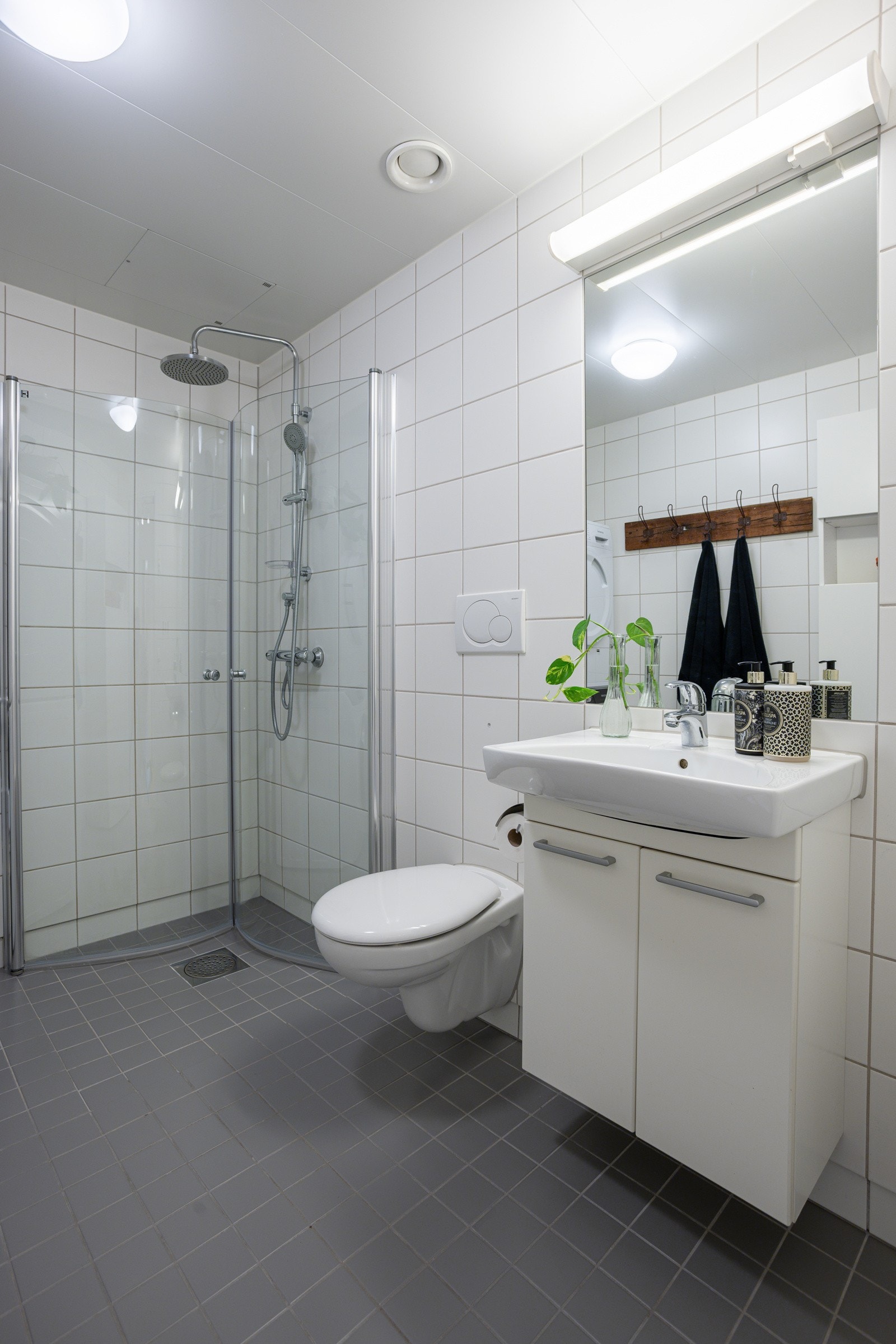 Tidløs baderomsinnredning med lyse fronter og ovenpåliggende servant, speil med belysning over. Baderommet er også utstyrt med dusjhjørne med innfellbare glassdører og både hånd- og regnfallsdusj, samt vegghengt toalett. Galleribilde