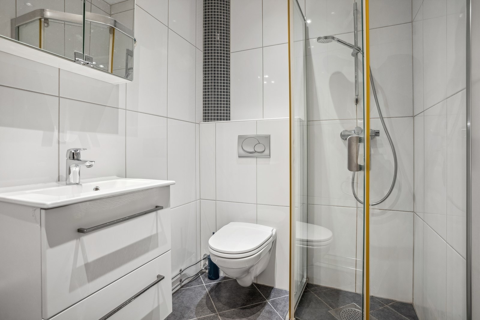 Badet er innredet med et dusjhjørne med glassdør, veggmontert toalett, en stilren innredning med heldekkende servant og speilskap med overliggende belysning. Galleribilde