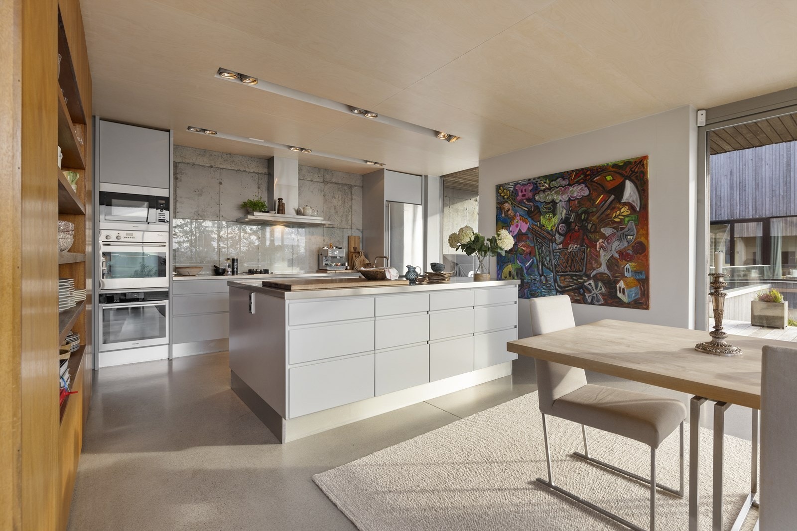 Stilren kjøkkeninnredning fra Boform bestående av glatte fronter med integrerte håndtak, stål benkeplater, integrert koketopp, stekeovner og oppvaskmaskin. Høykvalitets hvitevarer fra Míele og Gaggenau Galleribilde