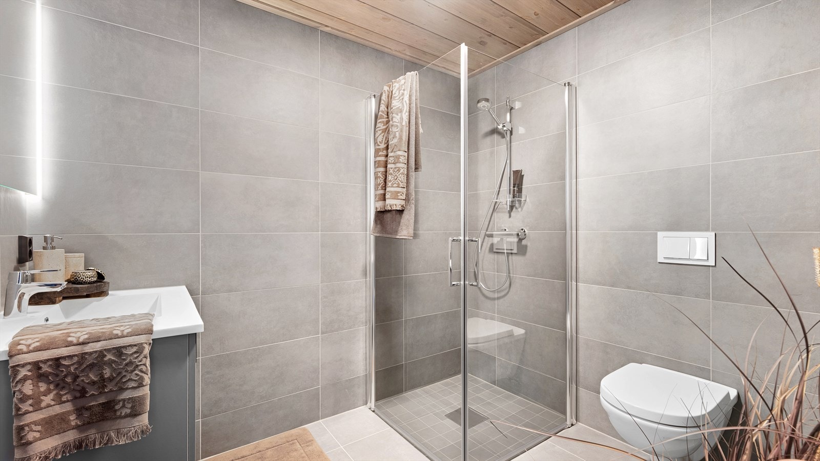 Baderommet inneholder et vegghengt toalett og dusjhjørne med dører i glass. Servantskap med skap med lyse, glatte fronter og speil over. Galleribilde