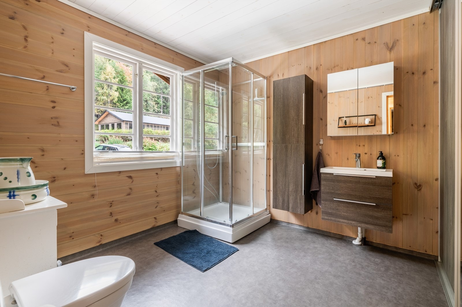 Romslig bad som innehar toalett, dusjkabinett og servantinnredning med underskuffer, sideskap og overskap med speildører. Det er opplegg til vaskemaskin. Galleribilde