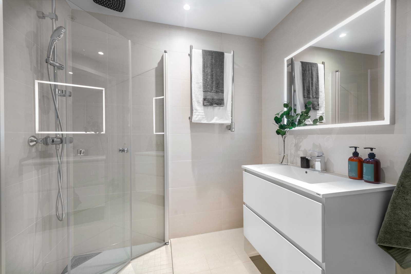 Badet med inngang fra entreen har servantskap med heldekkende servant og glatte fronter, speil med belysning, håndkletørker på vegg og et veggmontert toalett. Dusjhjørnet har hånddusj, regnfallsdusj og innfellbare glassdører Galleribilde