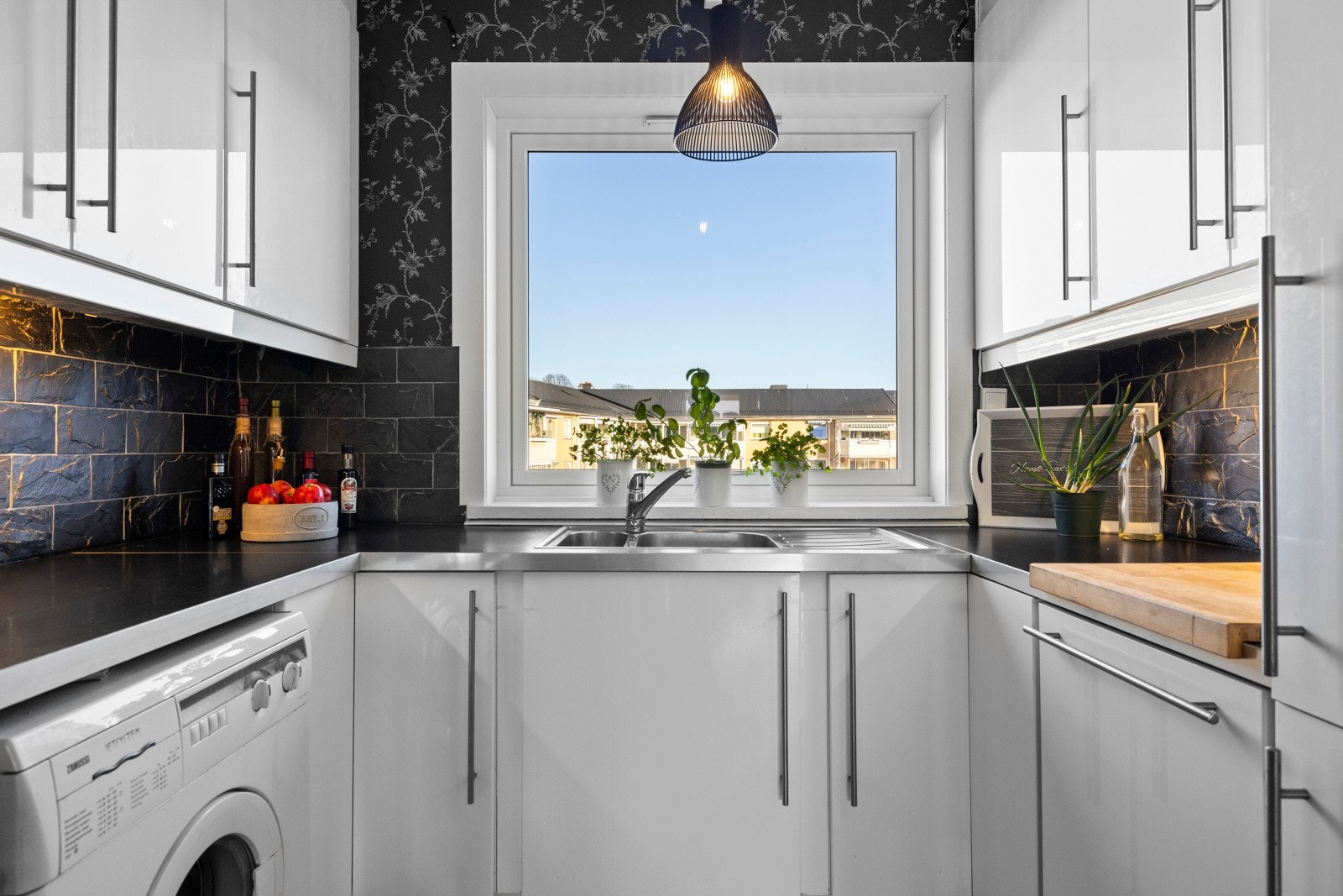 Nedfelt i benken, under et vindu, er det dobbel oppvaskkum med avrenningsfelt, og over er det sorte, rustikke fliser i mursteinsmønster og sort tapet Galleribilde