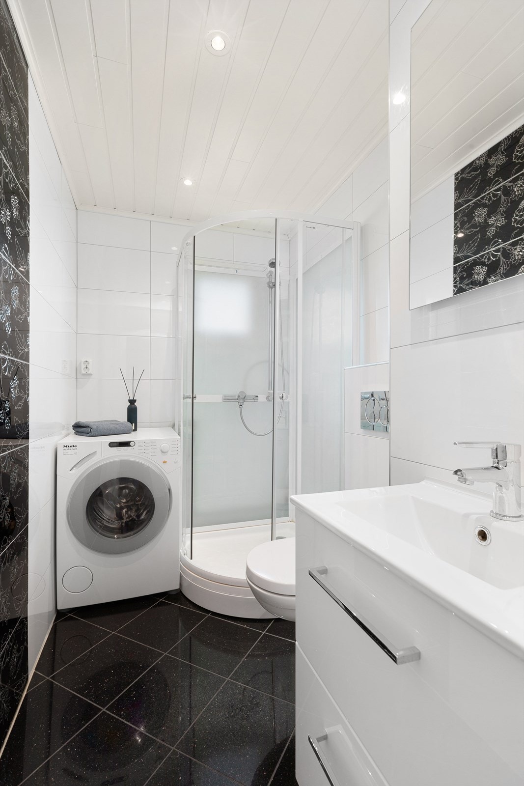 Pent flislagt bad med downlights i himling og innredning bestående av dusjkabinett, klosett og baderomsinnredning med speil. Badet har opplegg for vaskemaskin Galleribilde