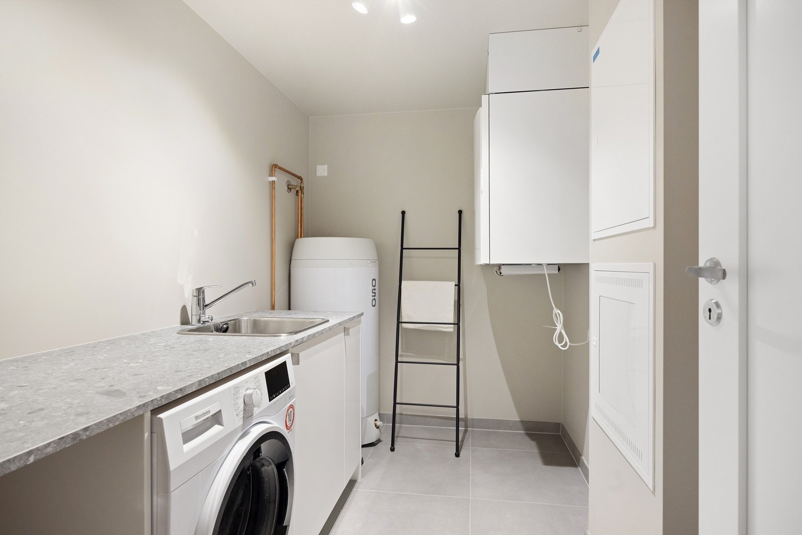 Praktisk vaskerom med malte plater på veggene og fliser på gulvet med elektrisk gulvvarme. Vaskerommet er innredet med skap og benkeplate med vask- og skyllekum, samt opplegg for vaskemaskin. Galleribilde