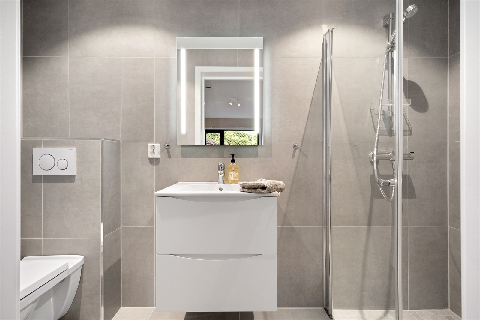 Delikat, flislagt baderom med innfelt downlights og elektrisk gulvvarme. Badet er innredet med vegghengt klosett, servant nedfelt i innredning, speilskap med lys og dusjhjørne med svingbare glassdører. Galleribilde