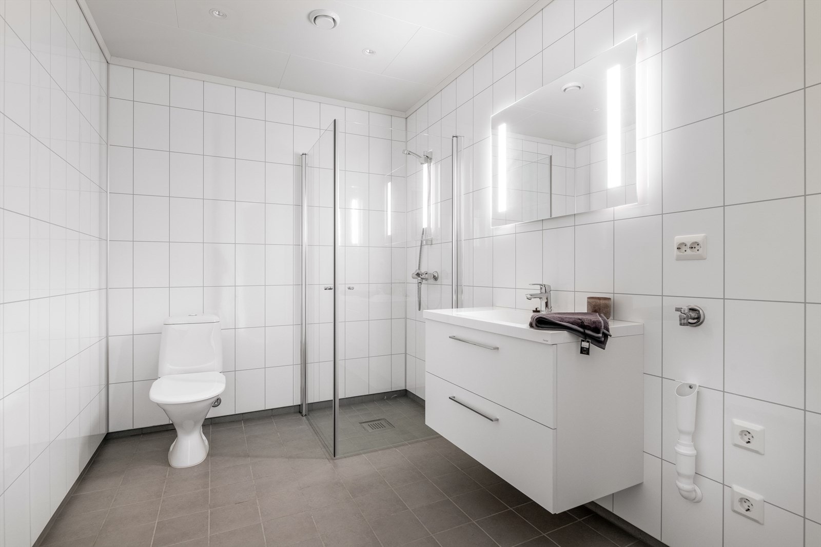 Lekkert baderom i boligen med flis på gulv med varmekabler. Pene baderomsplater på vegger og hvitmalte plater i himling med integrert belysning. Galleribilde