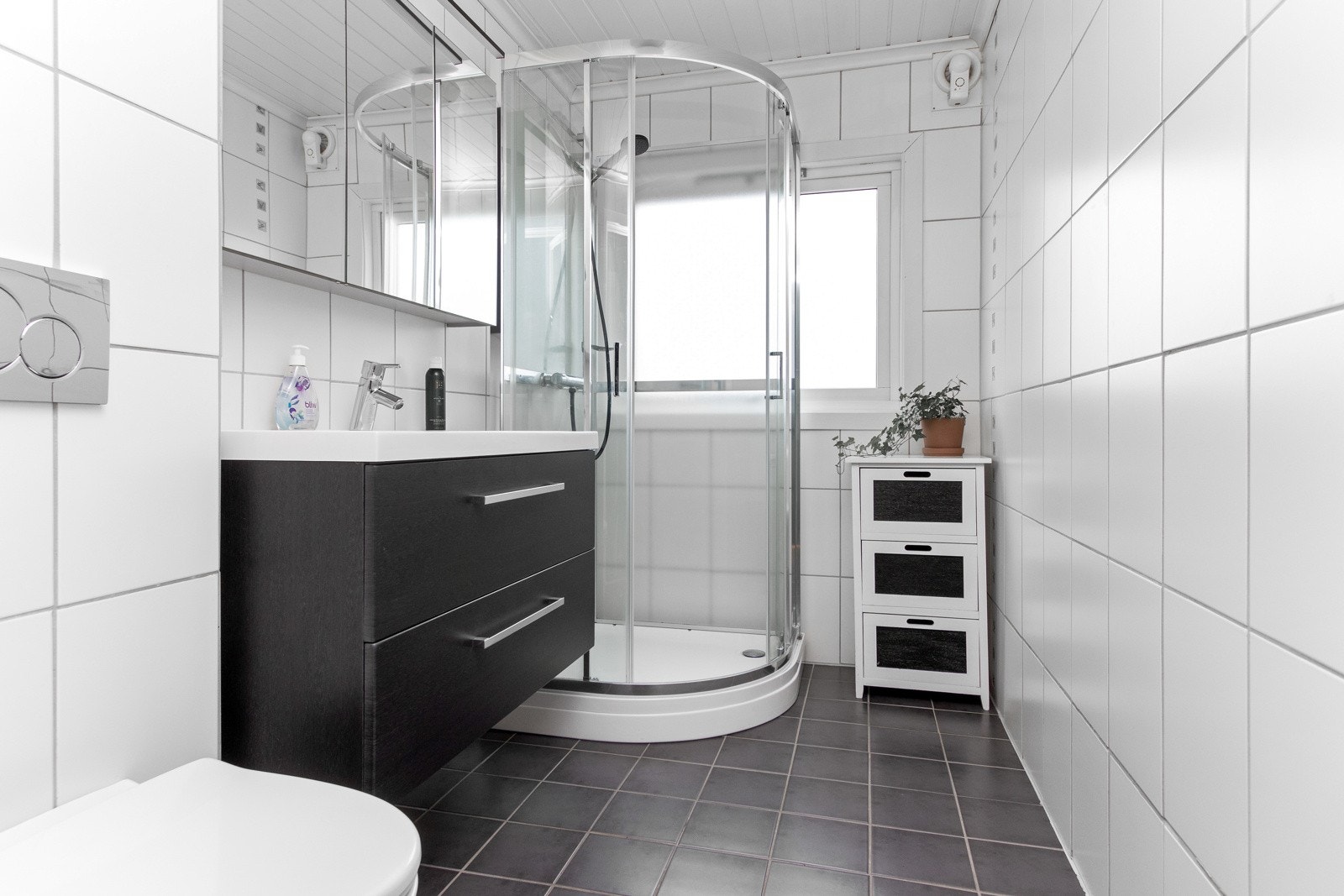 Badet har en nøytral stil med hvite fliser på veggene og sorte gulvfliser med gulvvarme. Videre er det innredet med vegghengt toalett, dusjkabinett og sort skuffeseksjon med heldekkende servant. Galleribilde