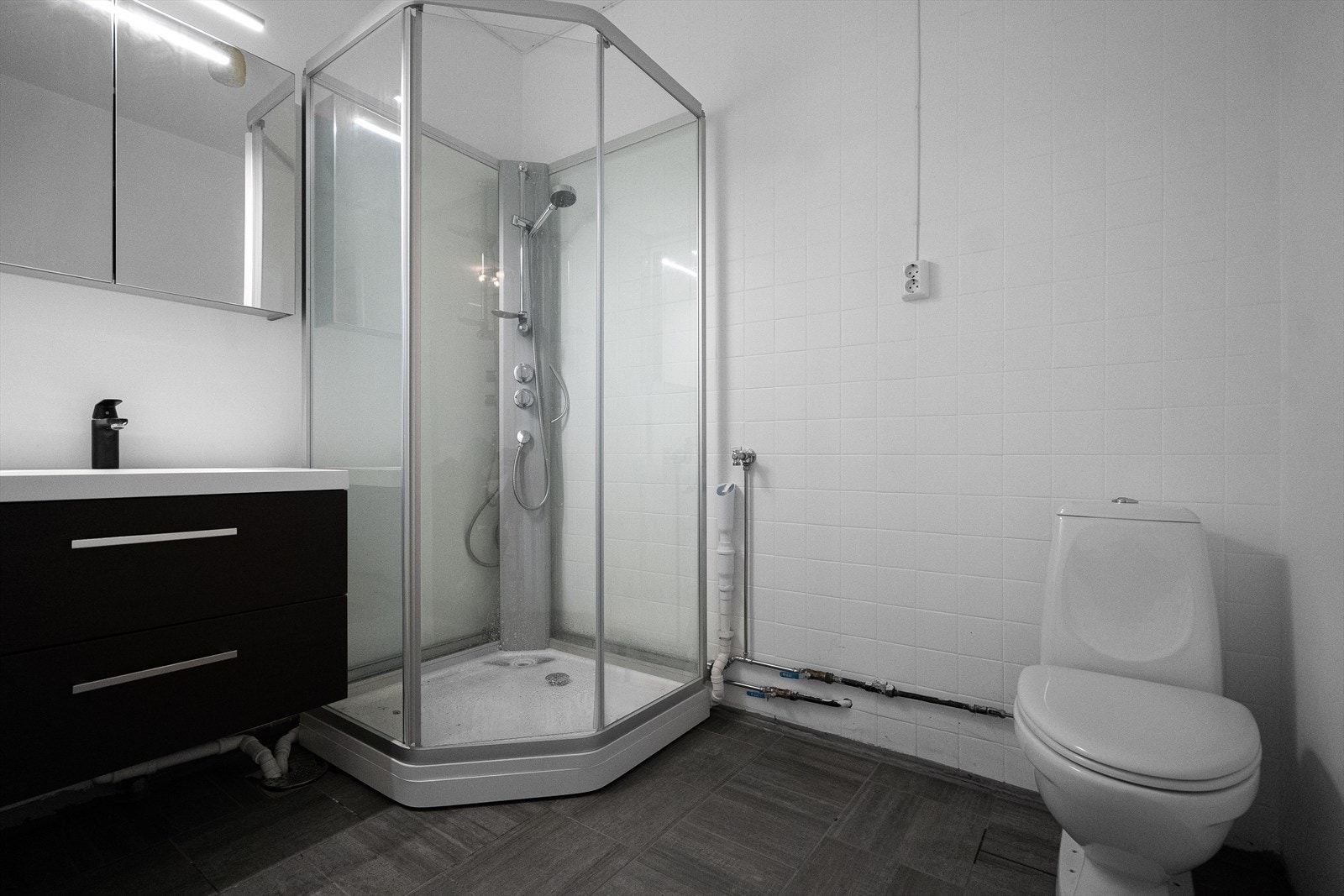 Baderommet har grå fliser på gulv med gulvvarme og hvite fliser på veggene. Det er plass og opplegg for vaskemaskin. Galleribilde