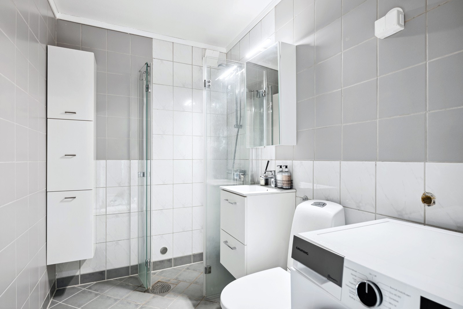 Flislagt bad med dusjplass med veggmonterte dører av glass, servant med over- og underskap, wc og vegghengt skap. Galleribilde