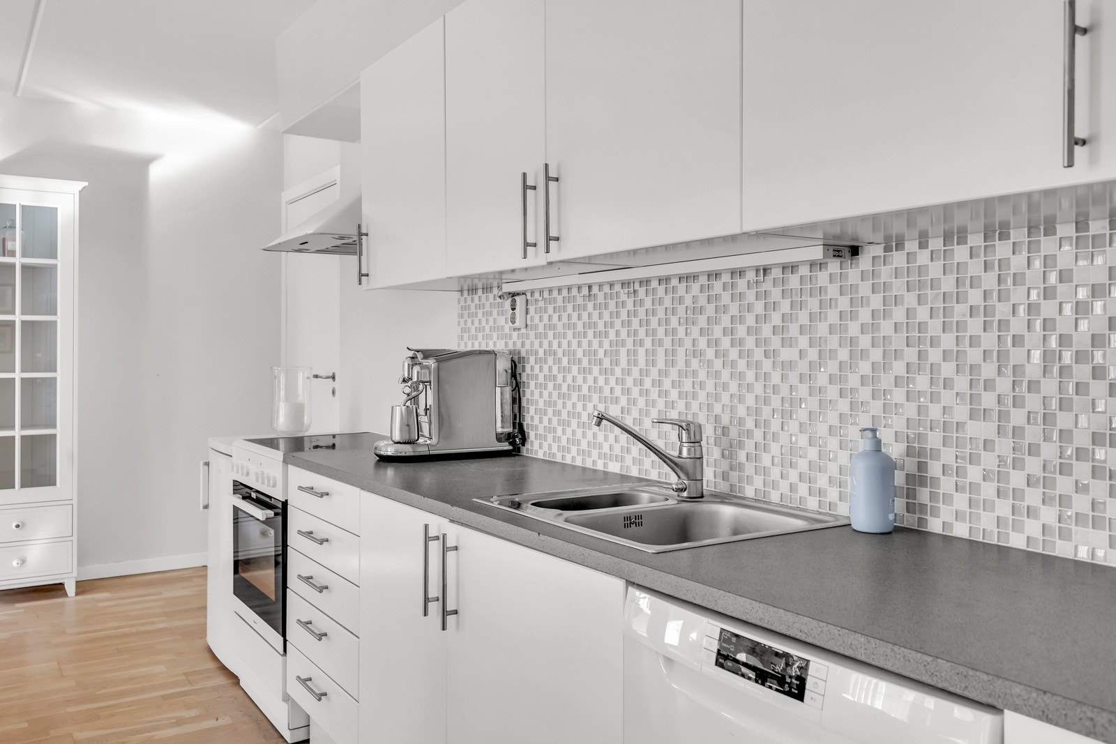 Kjøkkeninnredningen har glatte fronter med fliser på vegg over benkeskap og belysning under overskap. Det er oppvaskemaskin på kjøkkenet. Galleribilde