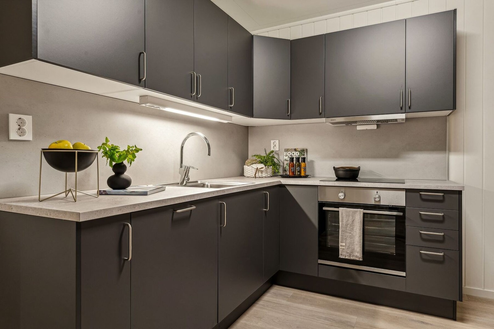 Vinklet kjøkkeninnredning med slette grå fronter og lys benkeplate av laminat. Integrert platetopp, stekeovn og oppvaskmaskin fra Electrolux. Ventilator fra Røroshetta. Galleribilde