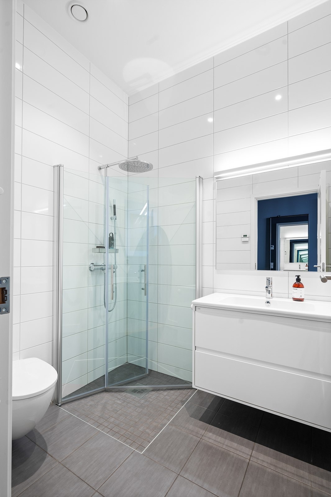 Boligens første baderom har tilgang fra hovedsoverommet og er delikat, med hvite fliser på vegg, grå fliser og varme i gulv samt downlights i tak Galleribilde