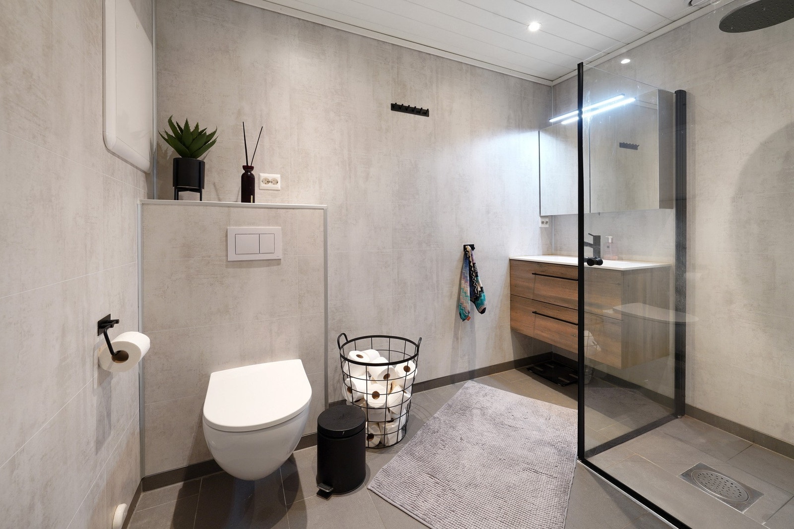 Moderne og stilrent bad med lyse fliser. Badet har en praktisk dusjnisje med glassvegg og vegghengt toalett Galleribilde